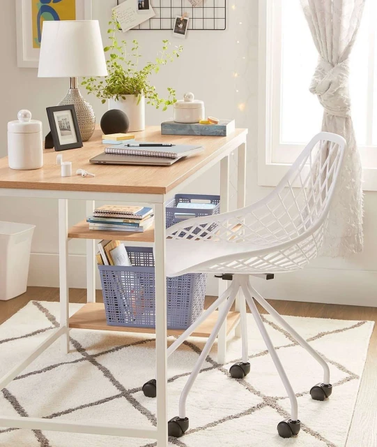 Home office setup with a light wood desk, white chair, lamp, and decor, set against a white and gray patterned rug and natural light.
