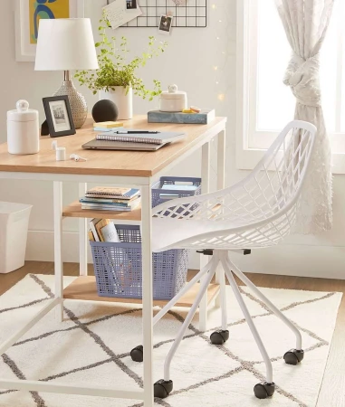 Home office setup with a light wood desk, white chair, lamp, and decor, set against a white and gray patterned rug and natural light.