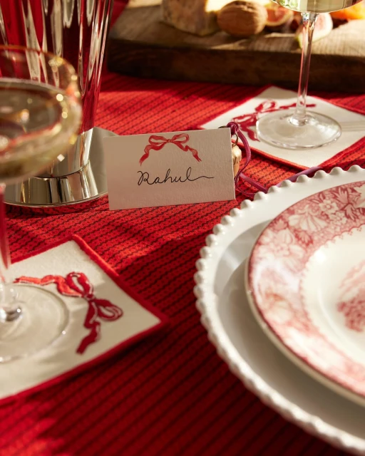 A table setting features a plate, crystal glass, name card with a bow illustration, and a red tablecloth, creating a festive and elegant ambiance.