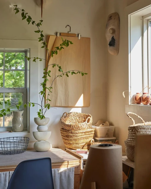 Sunlit interior photo of pottery vases, straw baskets, and foliage, set on a wooden table near a window with natural light. 