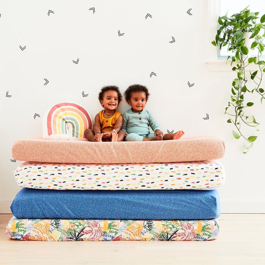 Two smiling babies in a room with decorative chevron wall decals sit on a stack of colorful, patterned textiles next to a rainbow pillow.