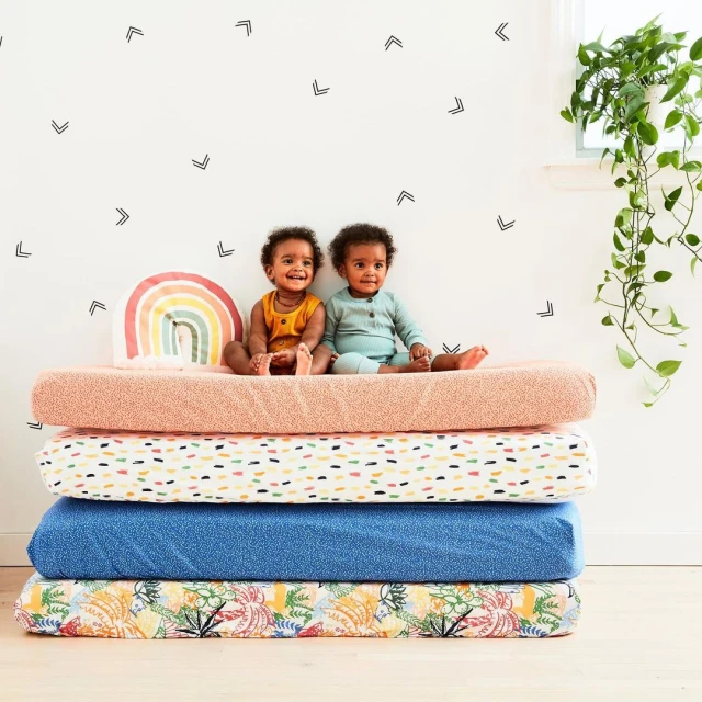 Two smiling babies in a room with decorative chevron wall decals sit on a stack of colorful, patterned textiles next to a rainbow pillow.