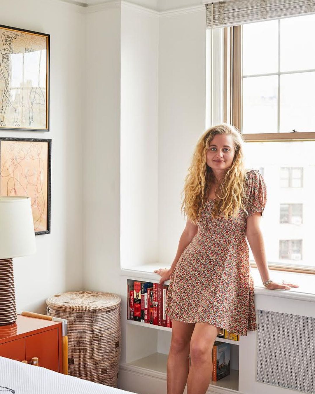 Adult woman stands in a room with a window, wearing a short, patterned dress; interior includes bookshelf, art, and woven hamper.