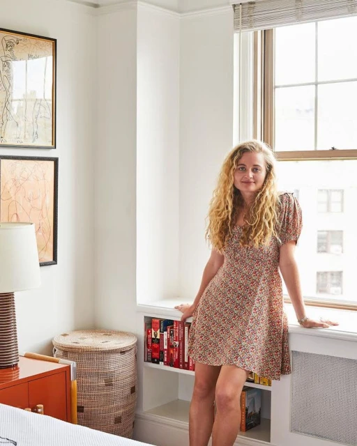 Adult woman stands in a room with a window, wearing a short, patterned dress; interior includes bookshelf, art, and woven hamper.
