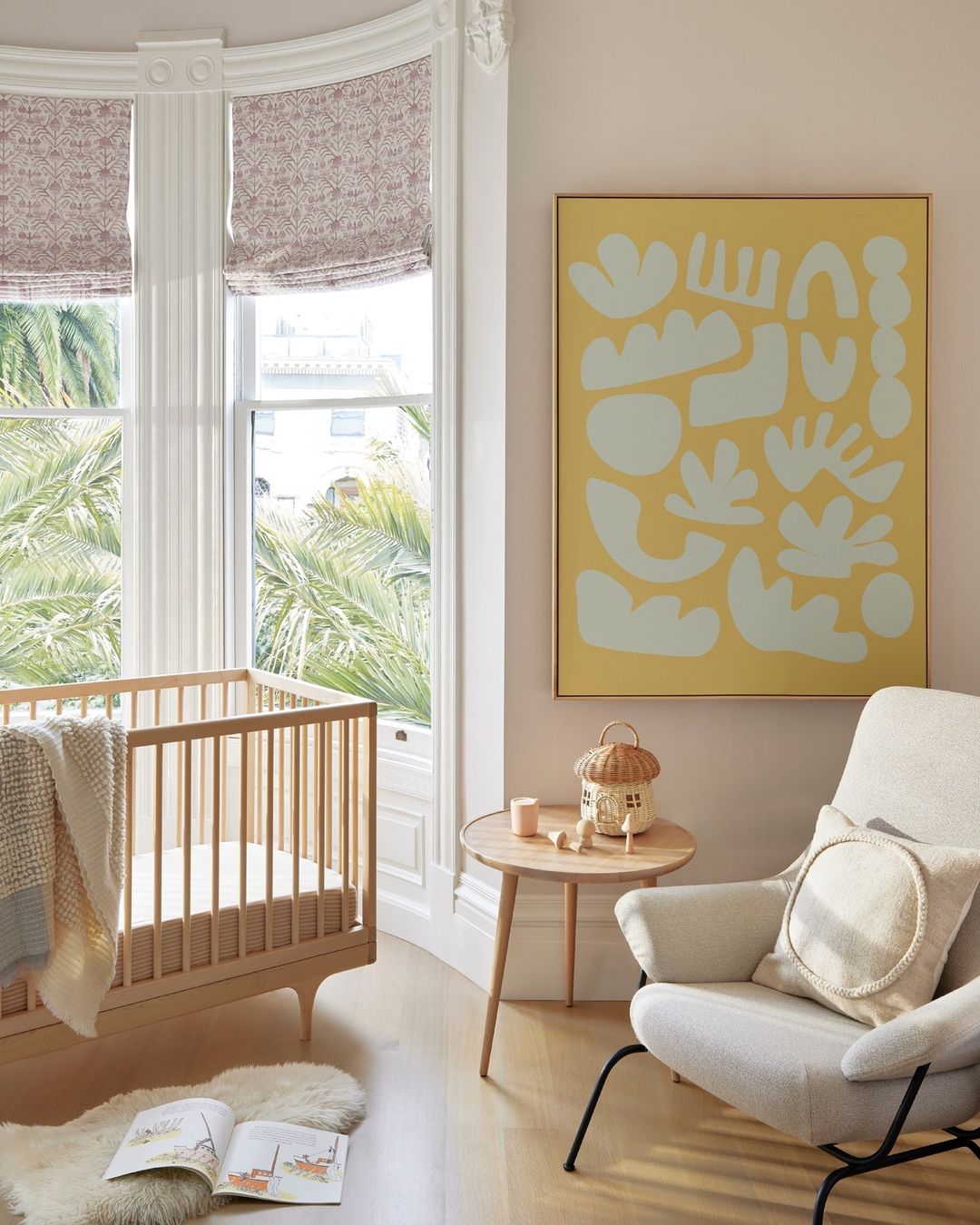 Interior of a child's room with a wooden crib, chair, side table holding decorations, abstract art, and large window providing natural light.