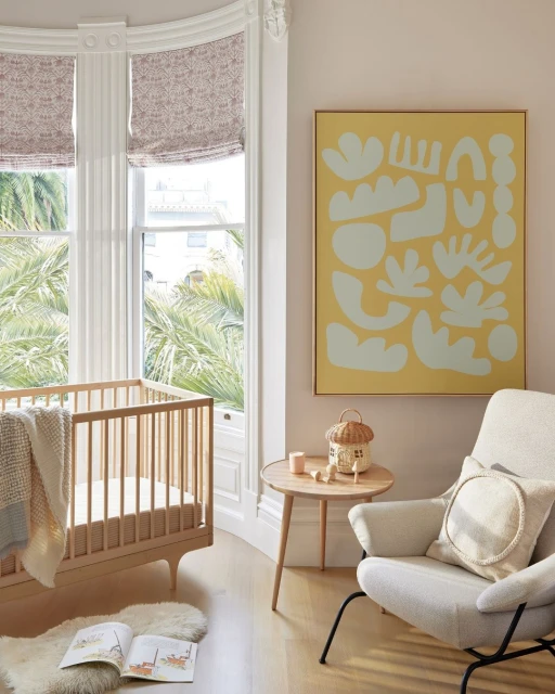 Interior of a child's room with a wooden crib, chair, side table holding decorations, abstract art, and large window providing natural light.