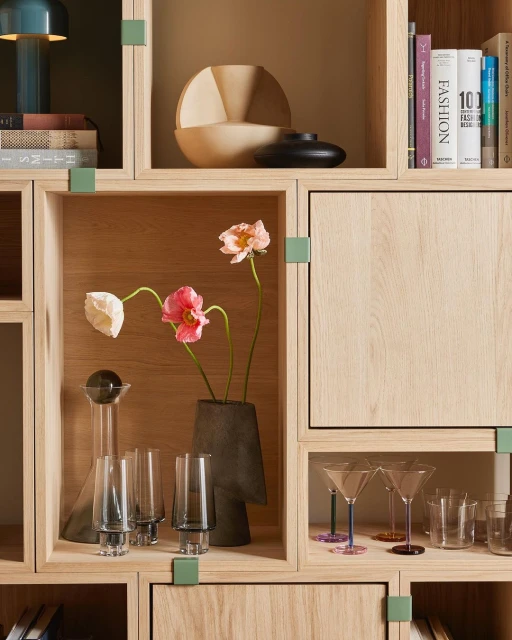 Interior view of wooden shelving unit featuring flower arrangements, decorative glass and ceramic vessels, and art and design books.
