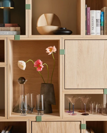 Interior view of wooden shelving unit featuring flower arrangements, decorative glass and ceramic vessels, and art and design books.