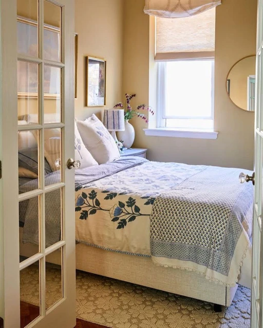 A cozy bedroom interior with a bed, nightstand, and window, softly lit and viewed through the opening of a pair of glass-paneled doors.