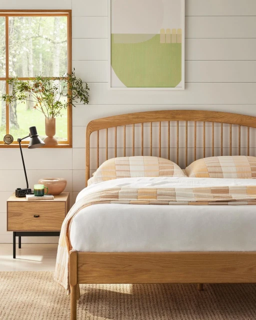 Bedroom interior with a wooden bed, a nightstand with a lamp, a framed artwork on the wall, and a window with a view of greenery.