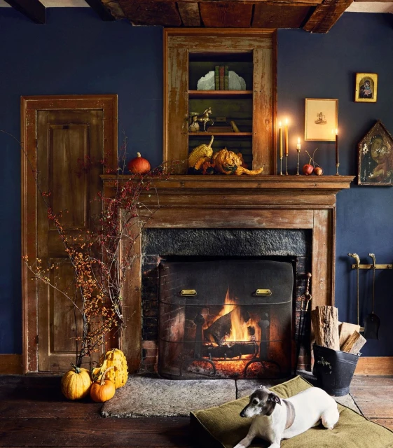A dog sits on a cushion in front of a fireplace with pumpkins, candles, and various decorative elements arranged above the mantelpiece.