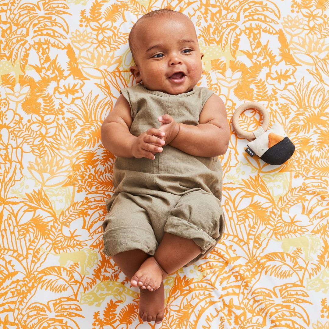 Happy baby in a light olive-green romper lying on a white blanket with a yellow floral and palm leaf pattern.