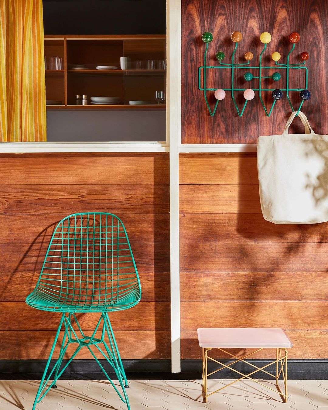 Interior view of a room with a teal wire chair, a small table, and a coat rack on a wood-paneled wall, showcasing modern design elements.