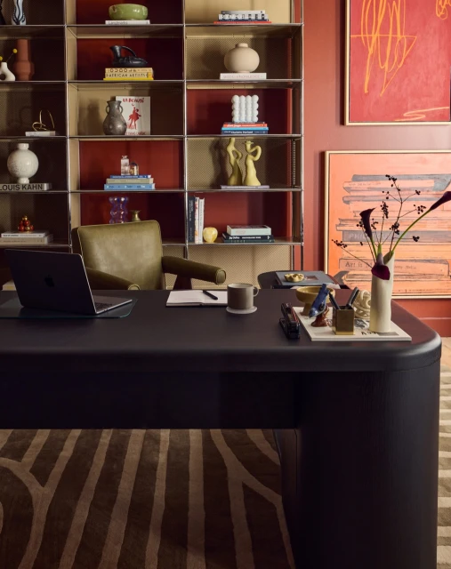 Home office featuring a black desk with a laptop, chair, bookshelf with various objects, and abstract art on the red walls.