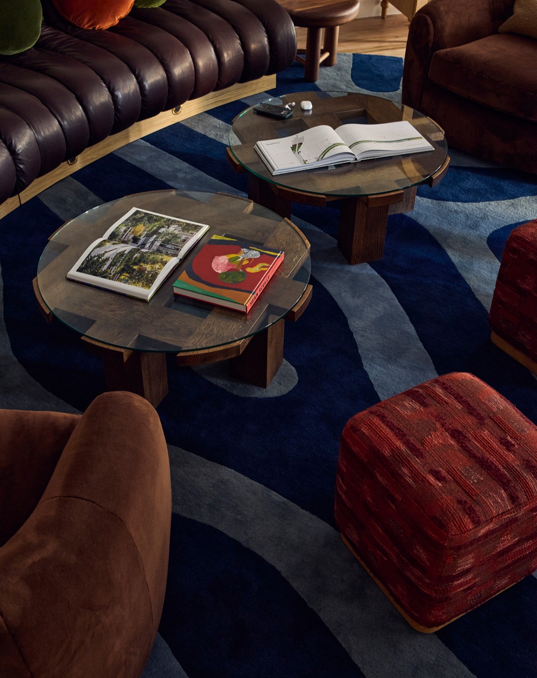 Interior design featuring a living room with a blue patterned rug, brown leather sofa, ottomans, armchairs, and round glass coffee tables.