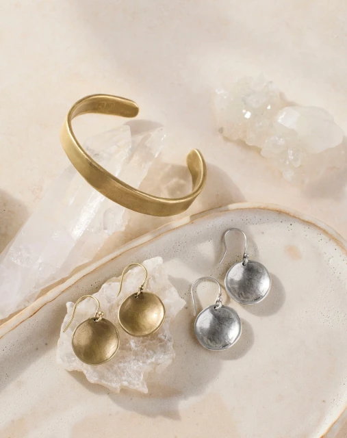 Close-up studio shot of gold cuff bracelet, with gold and silver earrings on a white decorative ceramic surface, soft lighting.