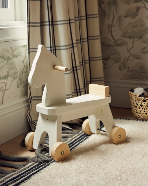 White wooden toy horse with wheels on a striped rug in a child's room with patterned wallpaper and curtains, providing a playful and cozy ambiance.