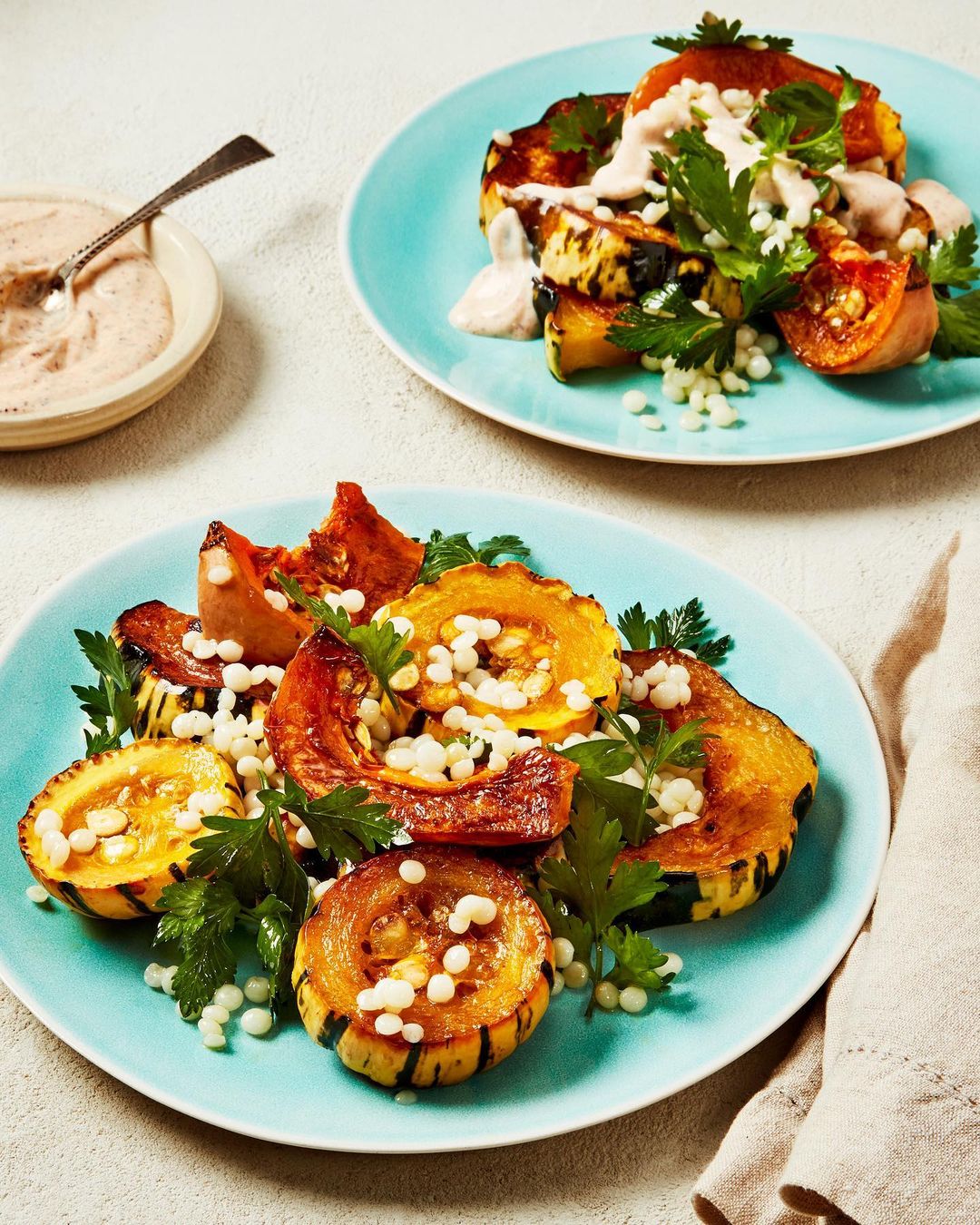 Roasted squash and couscous salad plated on blue plates with creamy sauce drizzle, set on neutral linen tablecloth.