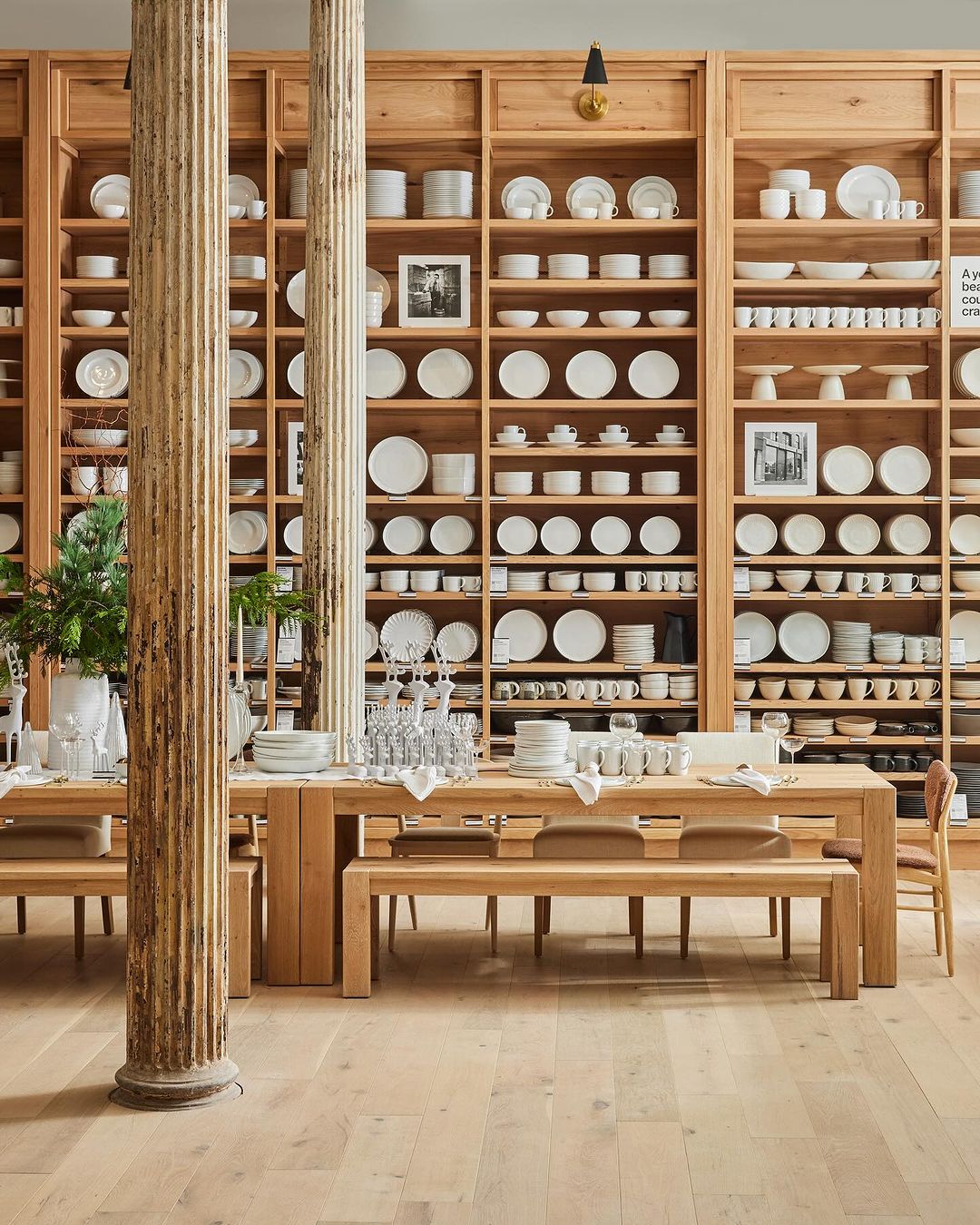 Interior view of a retail display featuring wooden tables, benches, and a large shelving unit stocked with white ceramic tableware.