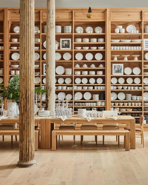 Interior view of a retail display featuring wooden tables, benches, and a large shelving unit stocked with white ceramic tableware.