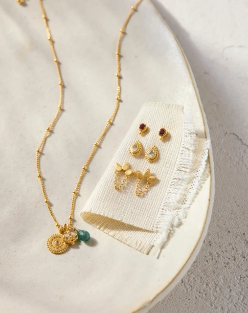 Overhead shot of gold beaded necklace with charms and earring sets displayed on a textured off-white surface, jewelry concept.