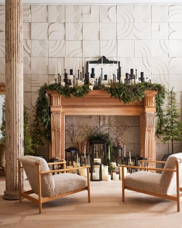 Two cream armchairs facing a wooden mantelpiece decorated with candles and greenery in an interior space
