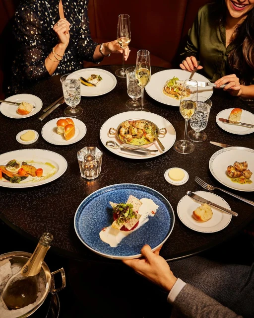 A group of people dine at a round table laden with gourmet dishes, wine glasses, cutlery, and a champagne bucket in a restaurant setting.