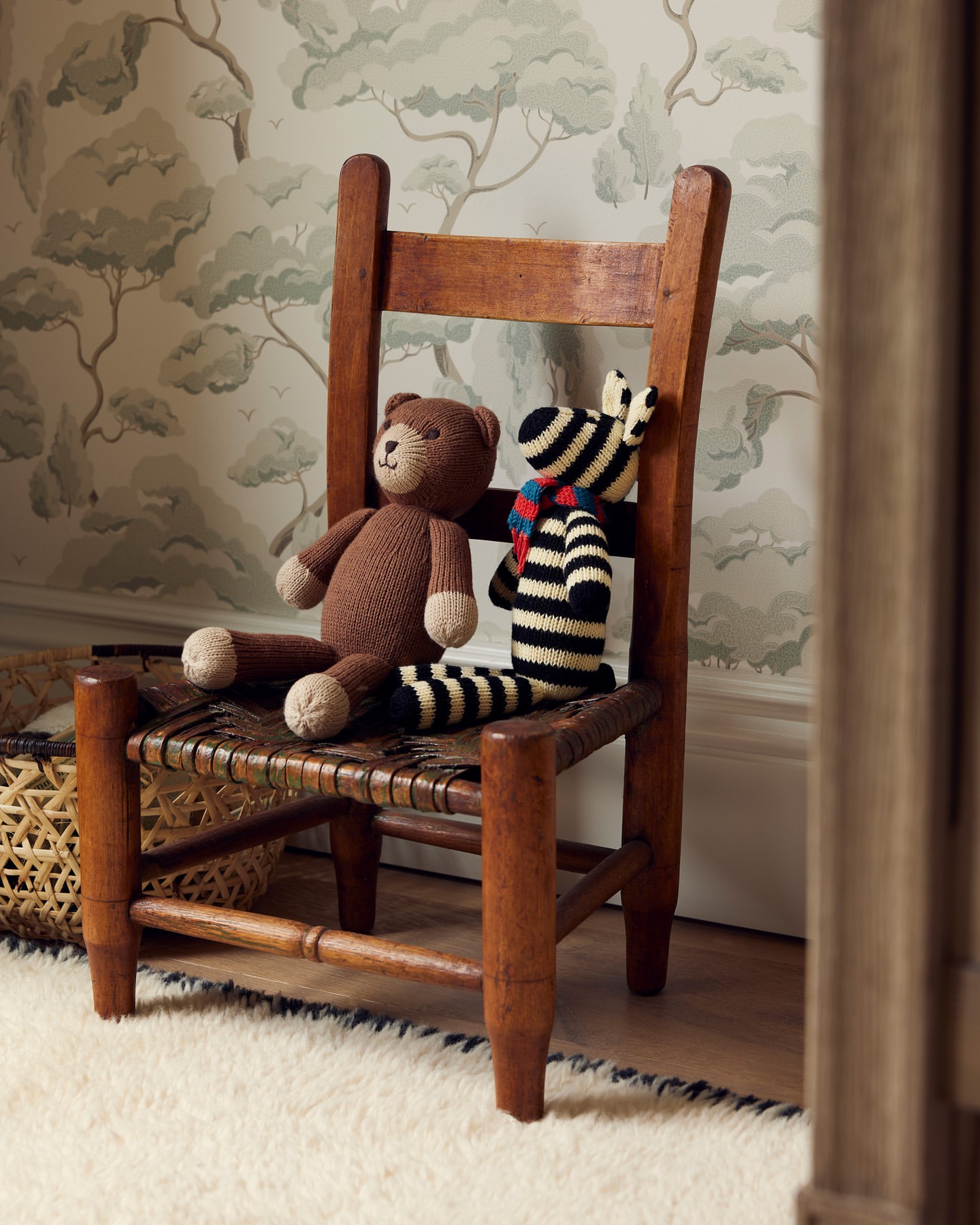 Stuffed bear and zebra toys sit on a woven-seat child's wooden chair, set against a wallpapered room with a tan and white rug.