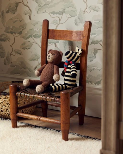 Stuffed bear and zebra toys sit on a woven-seat child's wooden chair, set against a wallpapered room with a tan and white rug.