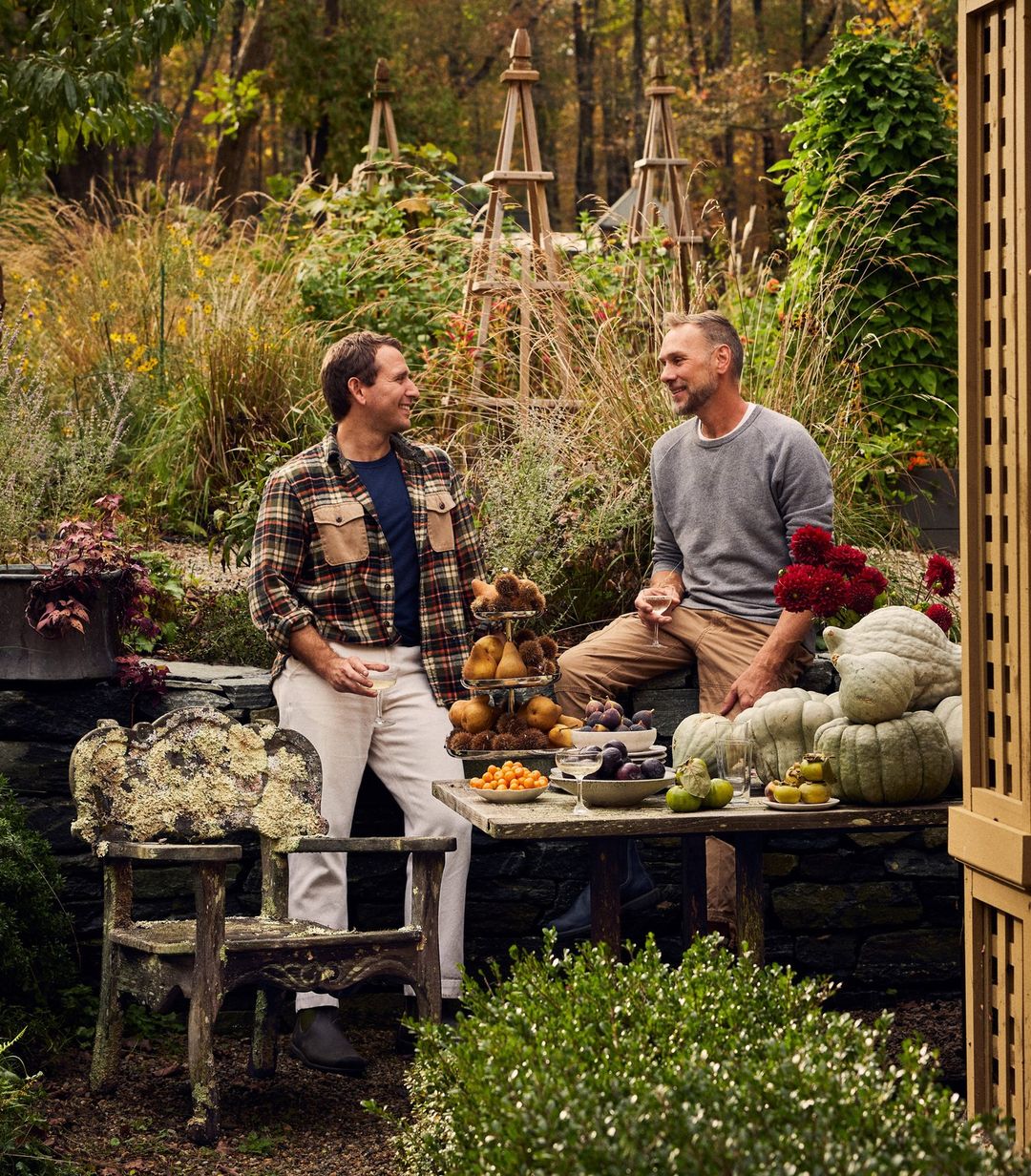 Two adult males are seated at a rustic table laden with fruit in a lush outdoor garden setting, featuring an aged wooden chair and stone wall.