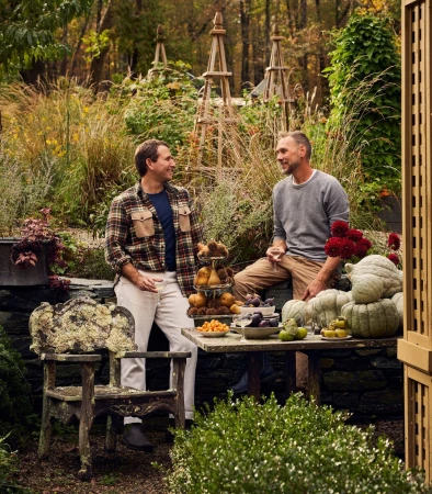 Two adult males are seated at a rustic table laden with fruit in a lush outdoor garden setting, featuring an aged wooden chair and stone wall.