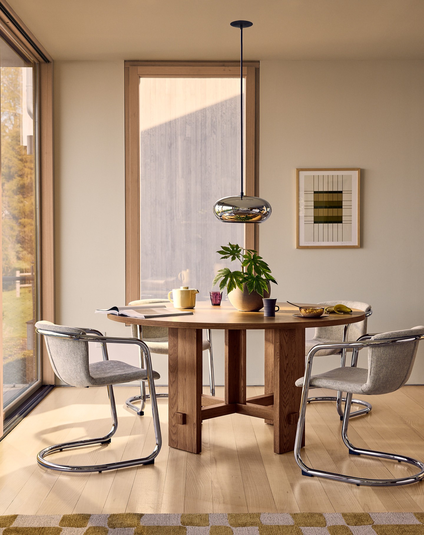 Round wooden dining table surrounded by chrome and fabric chairs in a bright modern interior with a pendant light