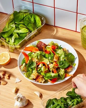 Overhead shot of a salad on a white plate with lettuce in plastic containers, lemons, garlic, and a hand placing leaves.