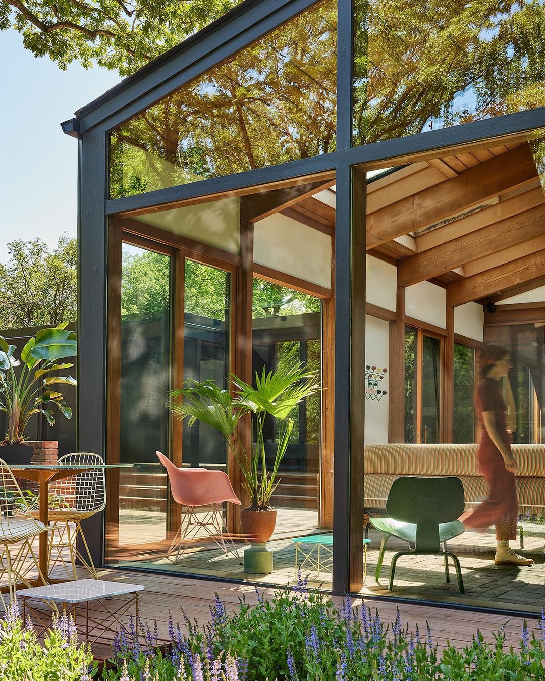 Modern glass-walled home with wood beams, surrounded by lush landscaping; dining area with a table, chairs, and potted plants.