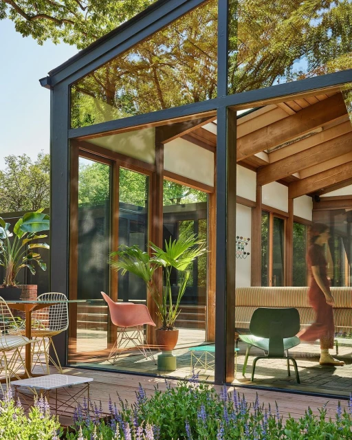 Modern glass-walled home with wood beams, surrounded by lush landscaping; dining area with a table, chairs, and potted plants.