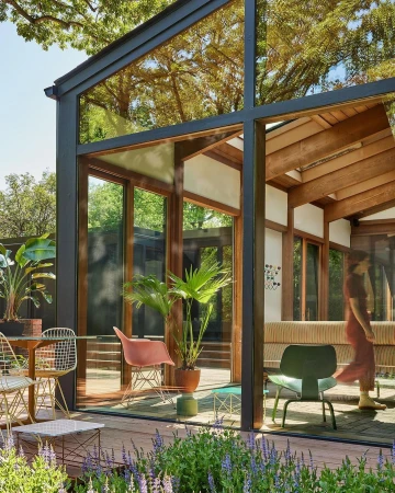 Modern glass-walled home with wood beams, surrounded by lush landscaping; dining area with a table, chairs, and potted plants.