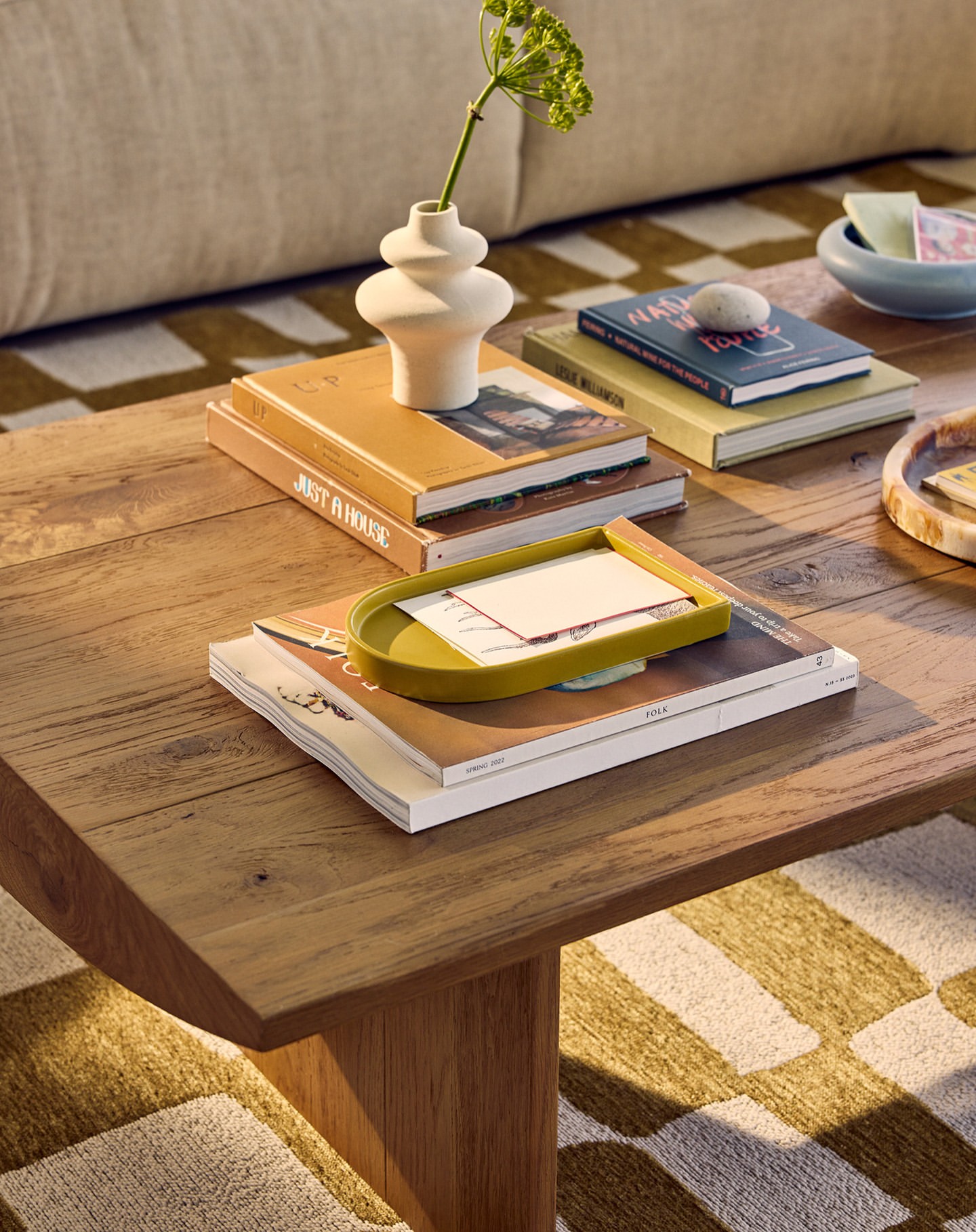 Still life on a wooden coffee table with books, a vase, and a green tray, set against a neutral-toned carpet with a geometric pattern.
