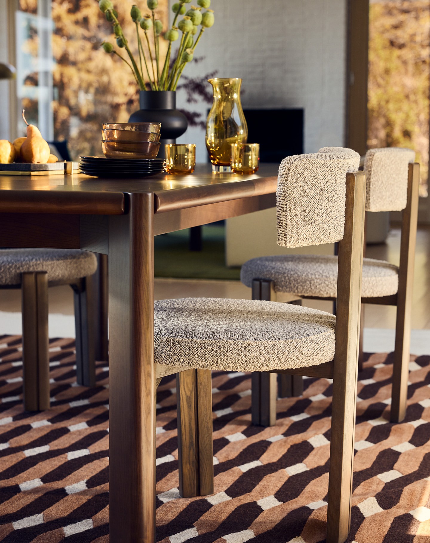 Dining table with upholstered chairs, set with dishes and glassware, under soft natural light with an abstract patterned rug.