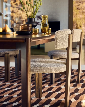 Dining table with upholstered chairs, set with dishes and glassware, under soft natural light with an abstract patterned rug.