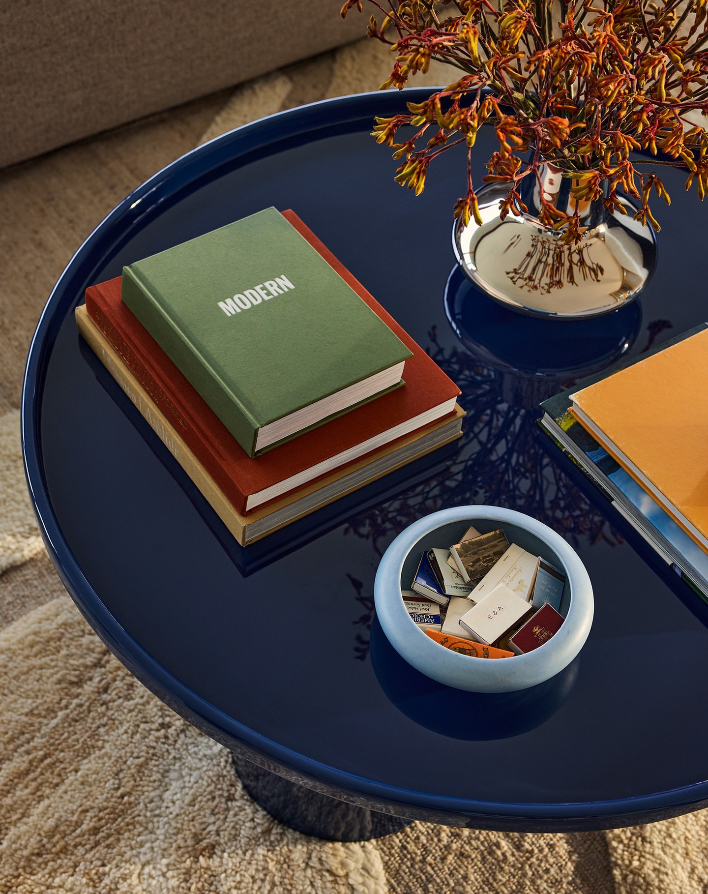 Close-up of a navy blue round table with books stacked with a green book on top with the word 'MODERN', a silver vase with flowers.