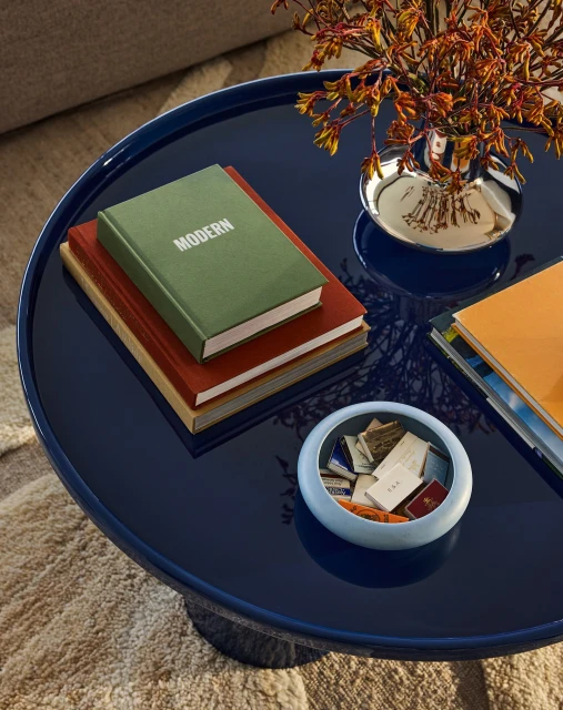 Close-up of a navy blue round table with books stacked with a green book on top with the word 'MODERN', a silver vase with flowers.