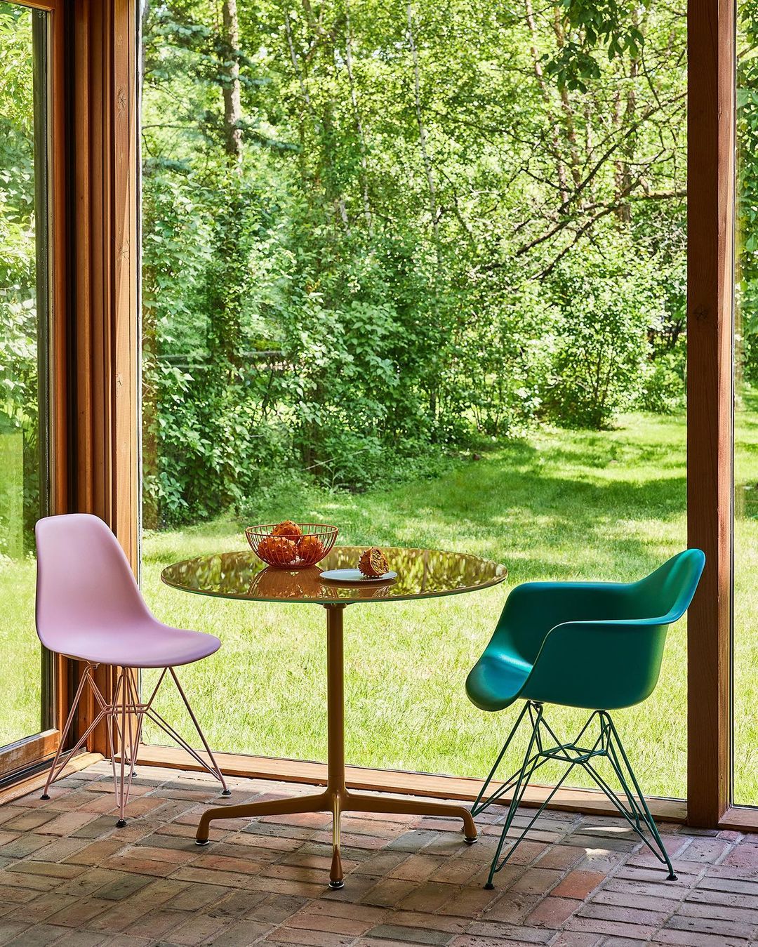 Indoor view of a round table with a bowl of fruit, a plate, and two chairs; one is pink and the other is turquoise.
