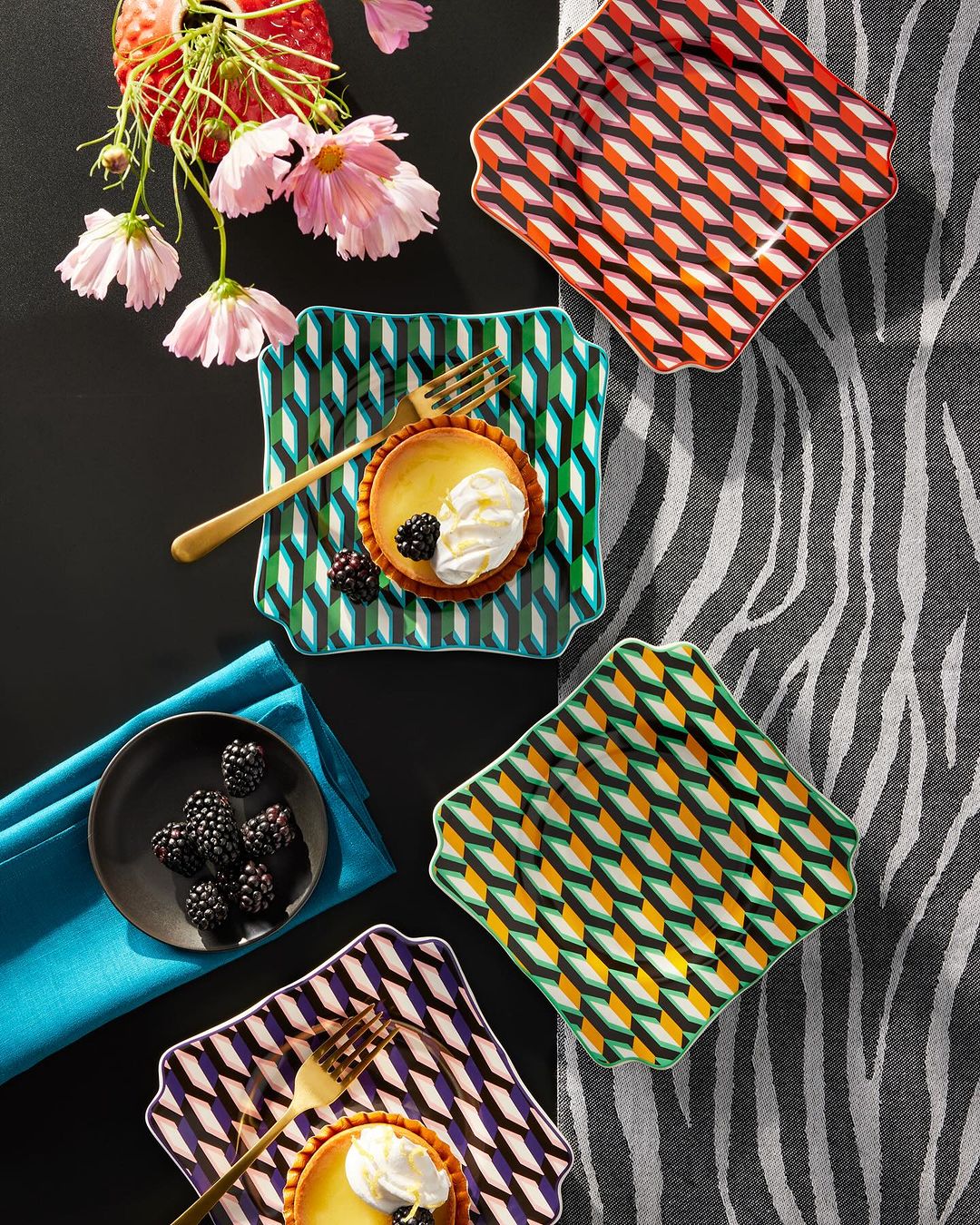 Overhead shot of patterned dessert plates, tarts with cream, blackberries, flowers and a gray zebra-striped background creates elegant food styling.