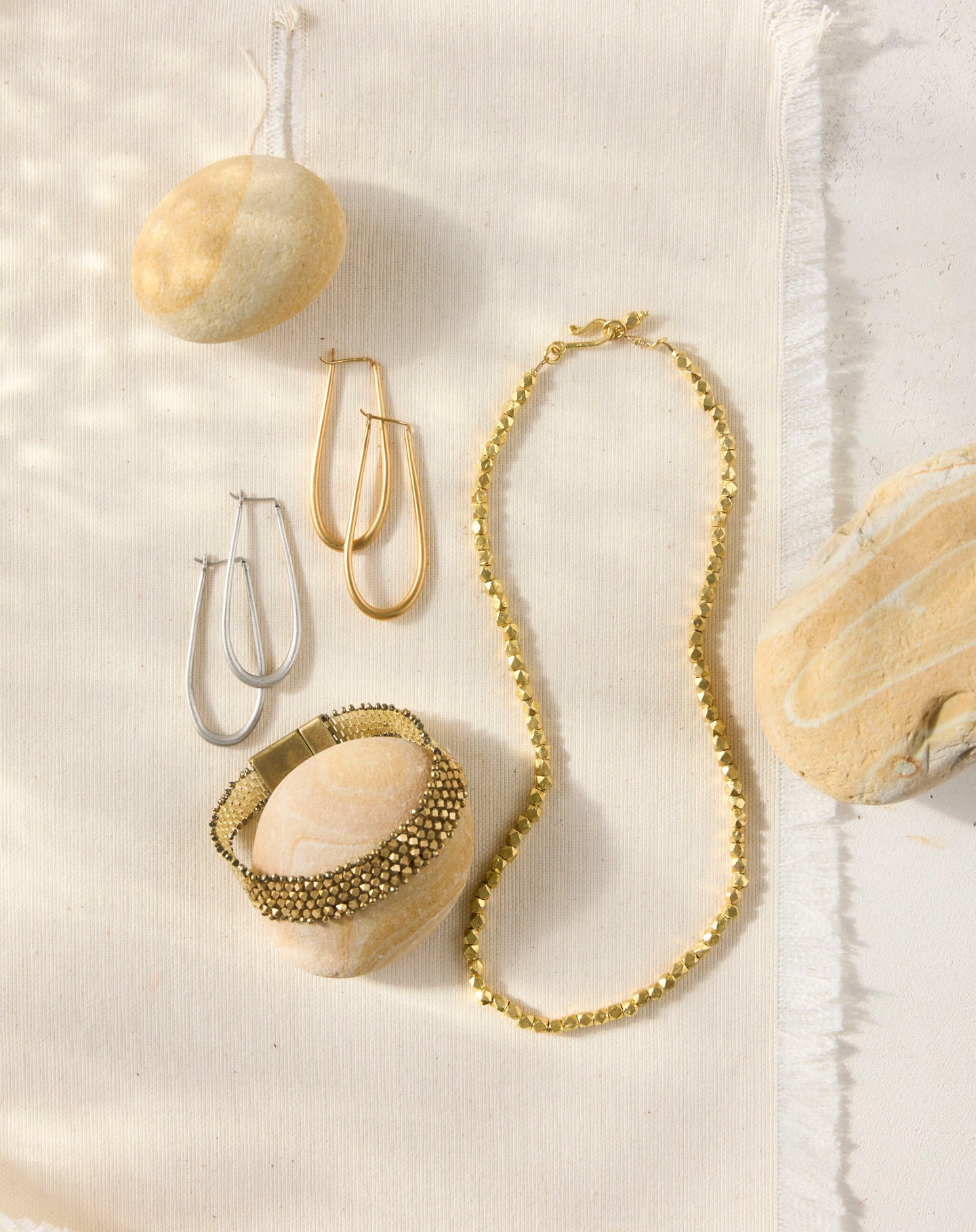 Still life of gold and silver hoop earrings, a gold beaded necklace, and a beaded bracelet displayed on neutral fabric and stones.
