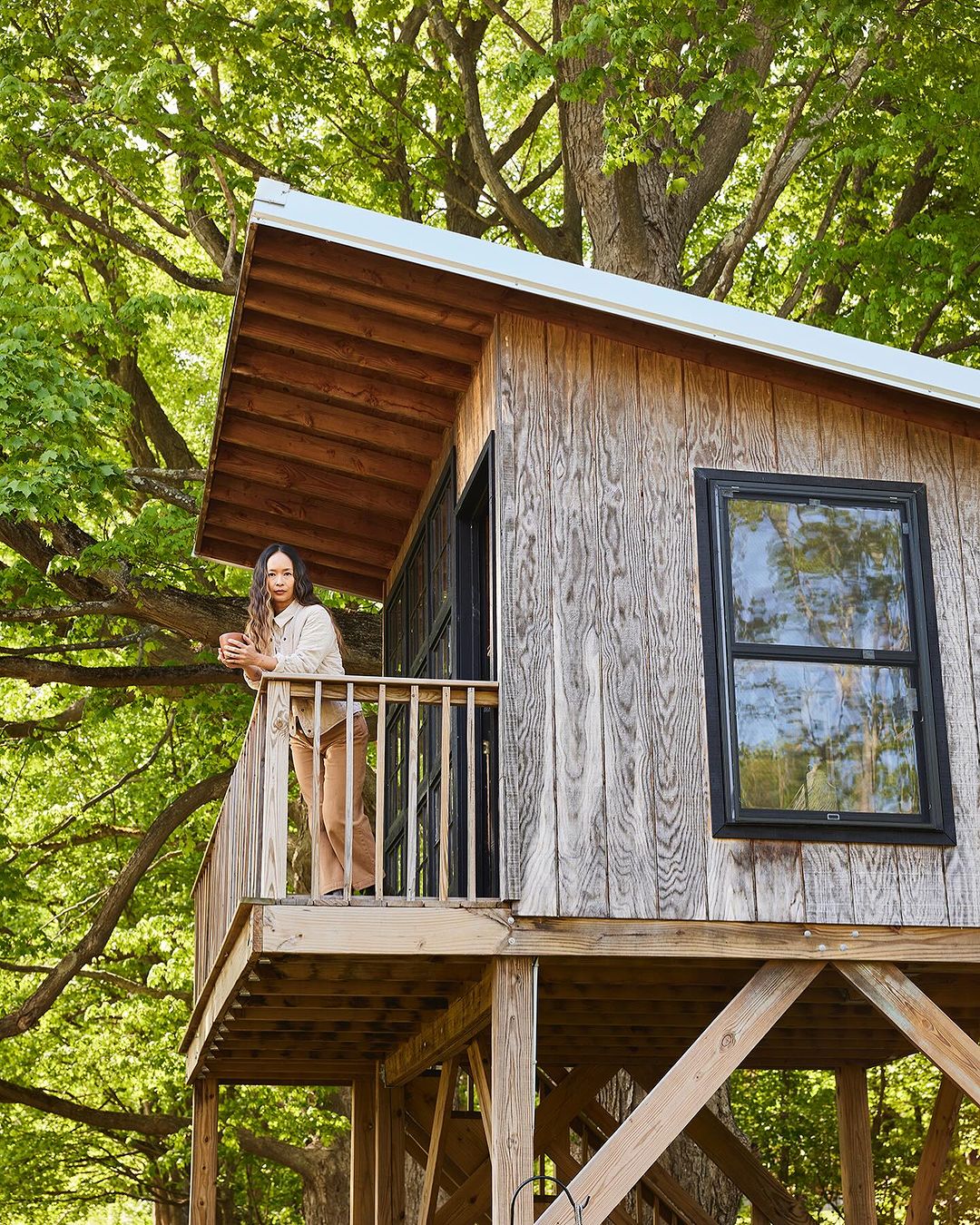 Adult woman stands on the wooden balcony of a treehouse surrounded by green trees and vegetation in a daylight outdoor setting.
