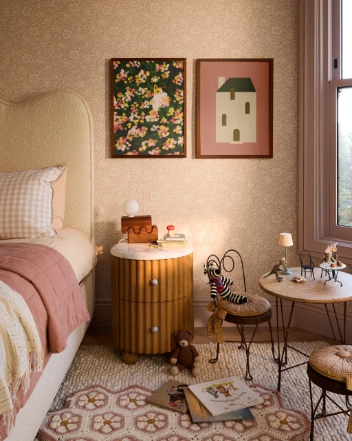 Bedroom interior with bed, nightstand, artwork, rug, chair, and children's books in a cozy, inviting space with soft, natural light.