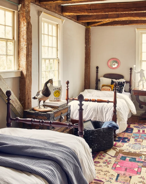 Bedroom interior with twin beds, wooden beams, goose figurine on trunk, and a colorful area rug creating a cozy, vintage aesthetic.