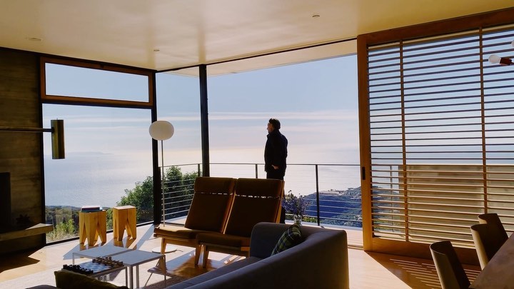 Man in black jacket looks out at ocean view from modern home interior with minimalist decor, including chairs, table, and large windows.