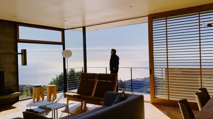 Man in black jacket looks out at ocean view from modern home interior with minimalist decor, including chairs, table, and large windows.