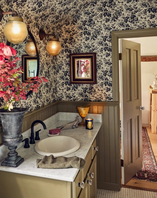 Bathroom interior with floral wallpaper, marble countertop, oval sink, red flowers in vase, and wall sconces providing warm lighting.